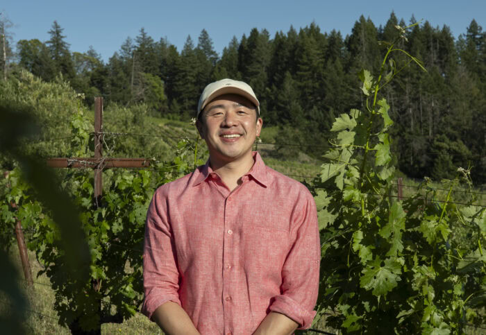 Smiling man in a pink shirt and white cap standing in a vineyard with green grapevines and trees in the background.